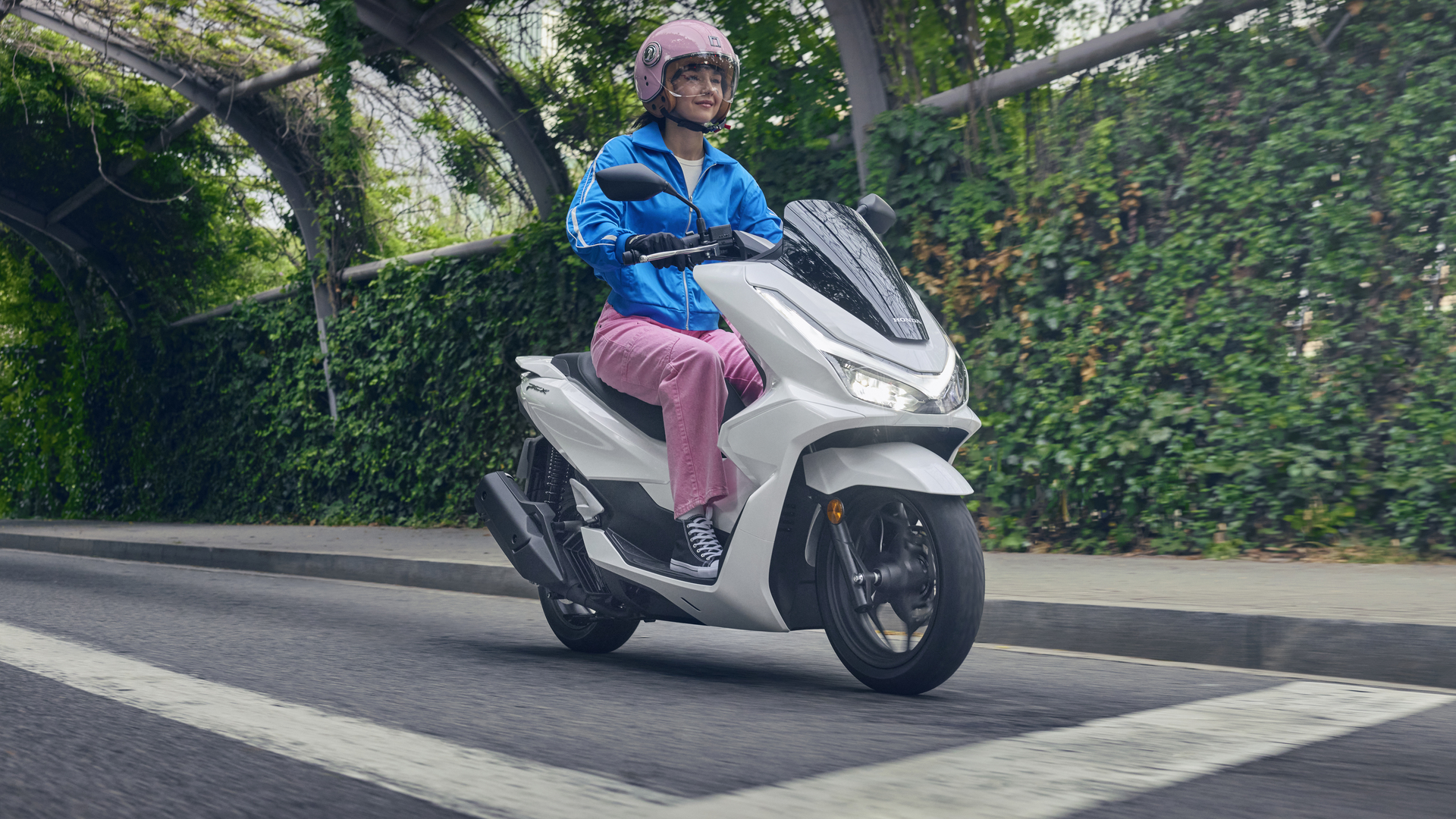 A person riding a white Honda PCX electric scooter on a tree-lined road. The rider is wearing a blue jacket, pink pants, and a helmet.