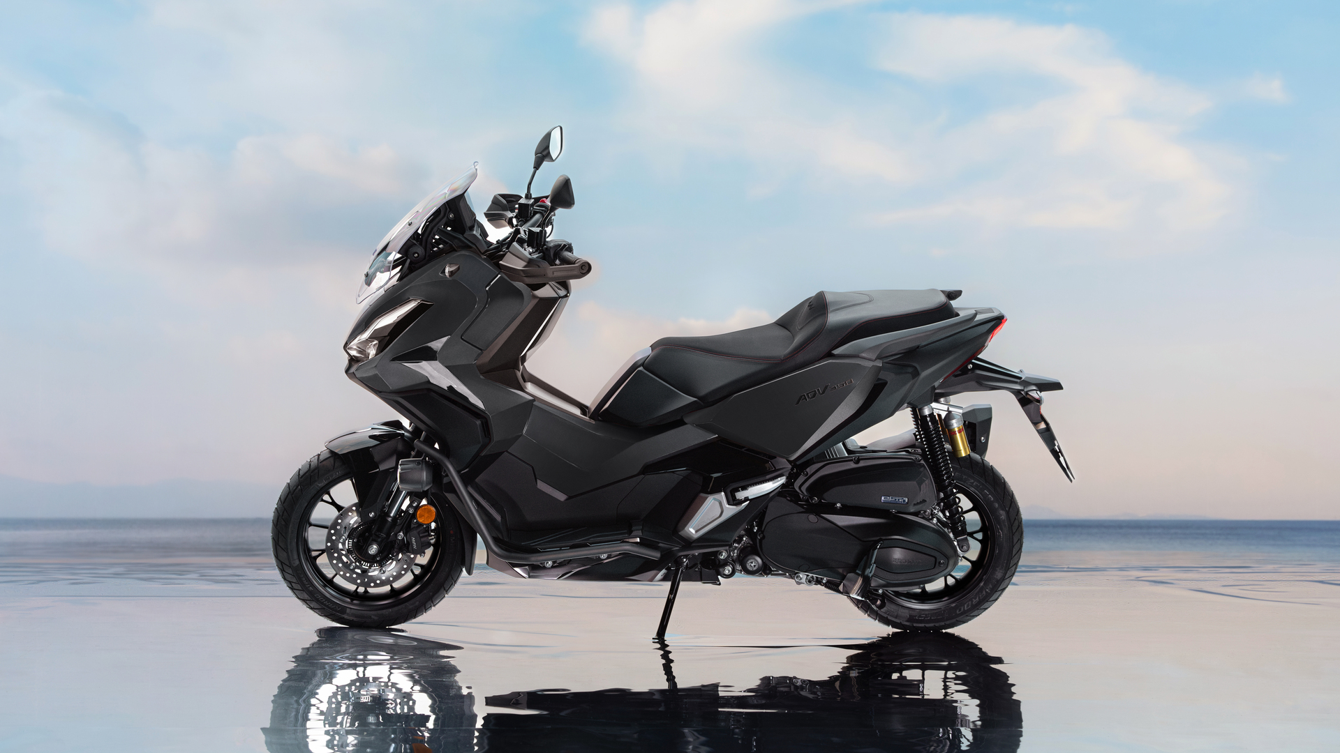A sleek black Honda Forza scooter parked on a reflective surface against a light blue sky background. The scooter's sporty design features a large windshield and aerodynamic body panels.