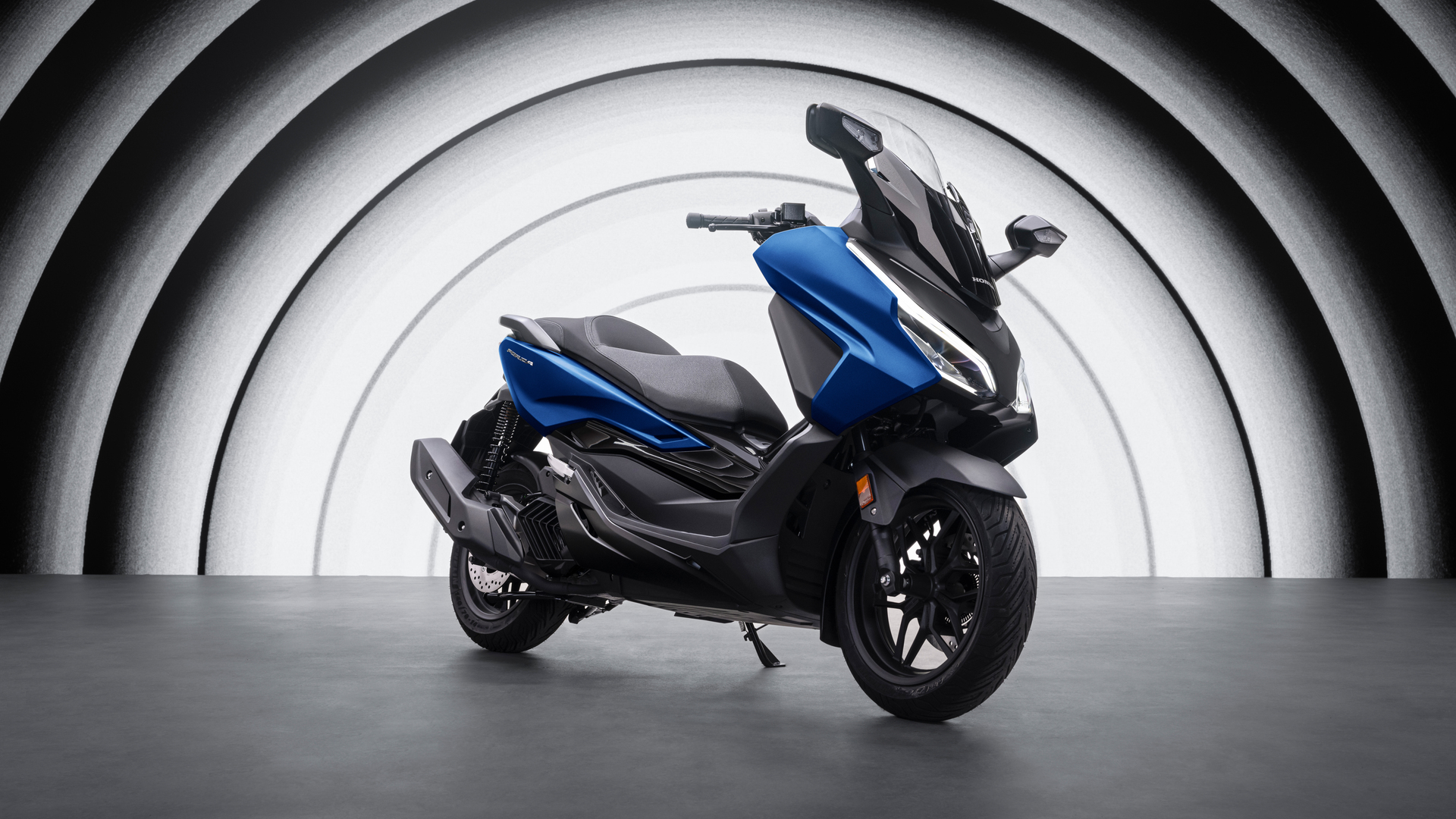A blue Honda Forza scooter positioned in the center of a circular gray and white backdrop. The sleek, modern scooter features a sporty design with a large windshield and aerodynamic body panels.
