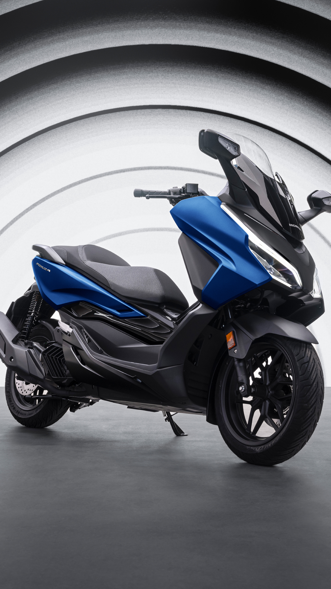 A blue and black Honda Forza scooter positioned against a background of concentric gray circles. The sleek, modern scooter features a large windshield and streamlined body design.