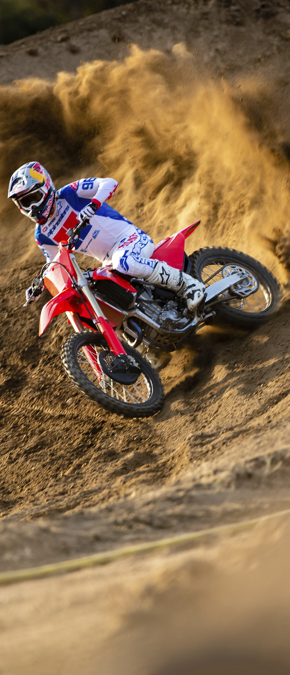 Un pilote de motocross en tenue blanche, bleue et rouge couche sa moto tout-terrain rouge dans un virage sablonneux relevé, projetant une grande gerbe de terre en pleine accélération.