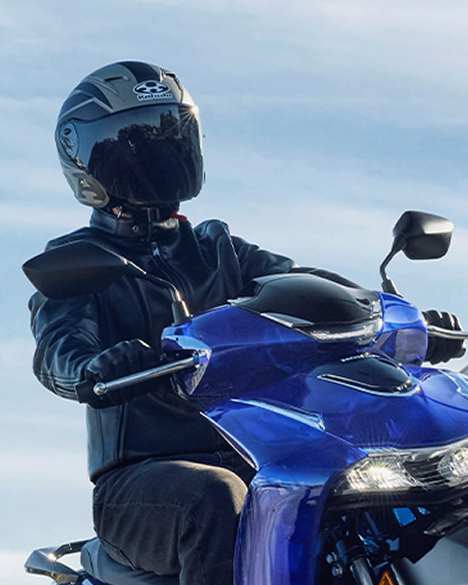 A motorcyclist wearing a black helmet and jacket sits on a blue Honda sport bike, likely a CBR model. The bike's front fairing and headlight are visible against a light blue sky background.