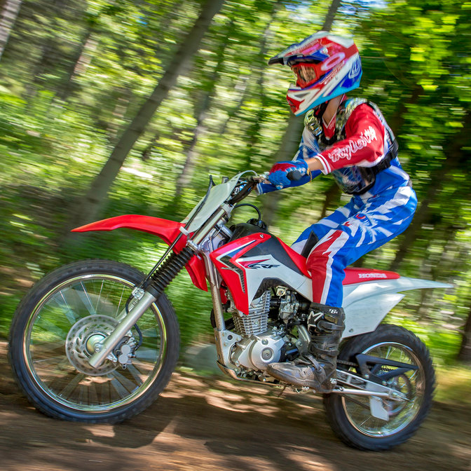 Vue latérale de la CRF125F avec conducteur sur chemin en terre, lors d'une course en forêt. gauche.